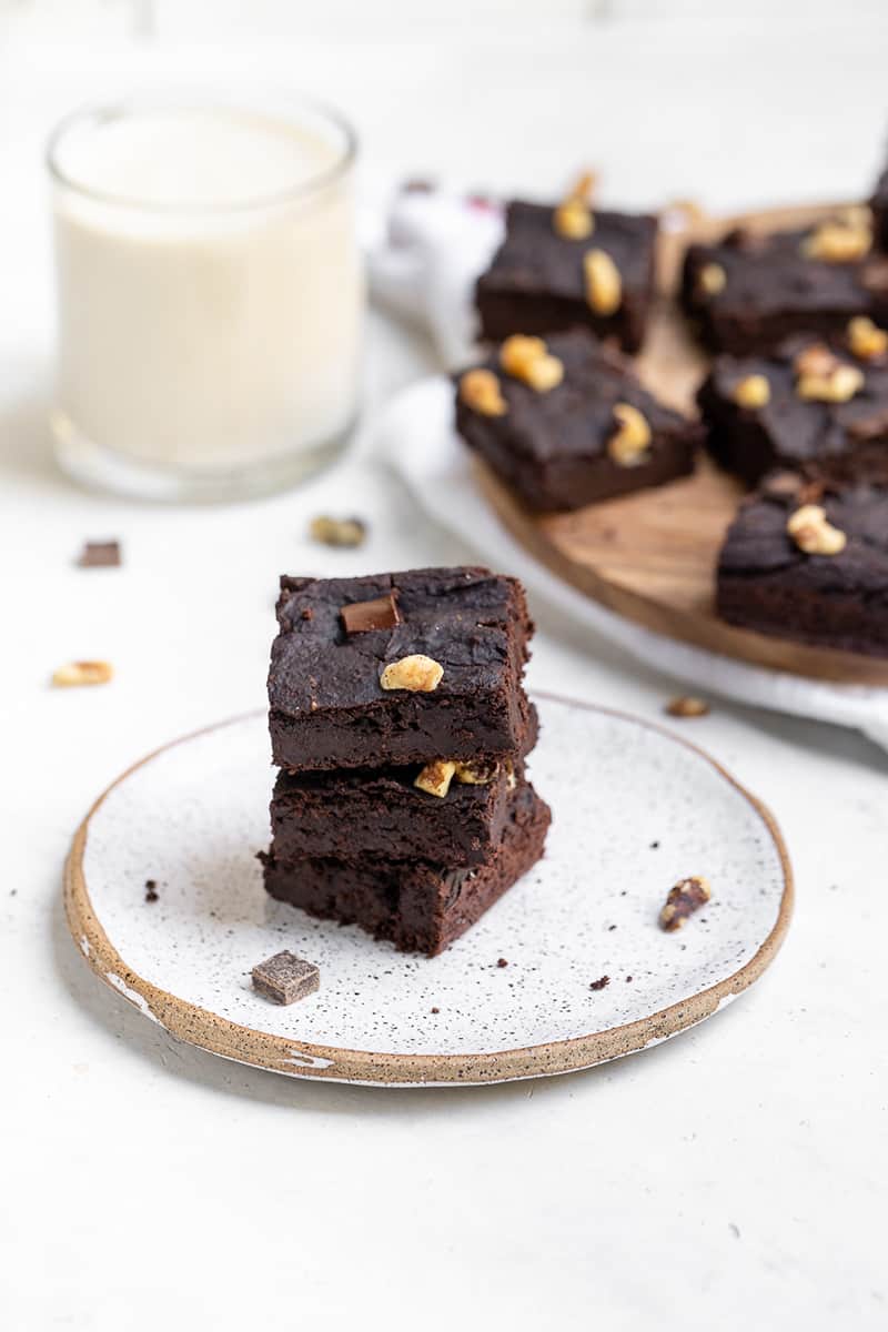 three black bean brownies stacked on a white plate in front of a plate of brownies