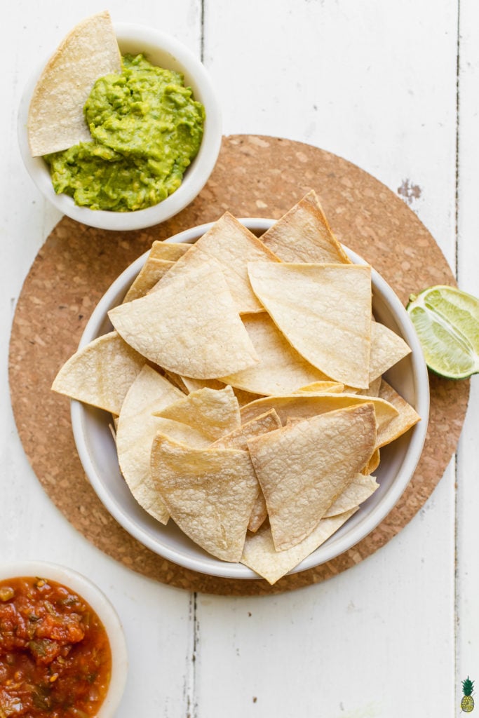 Homemade tortilla chips in a bowl on a cork board with guacamole, salsa and lime