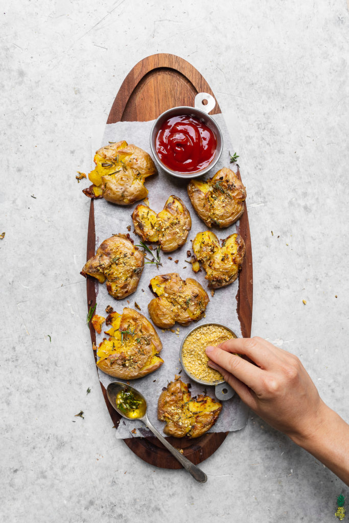 hand trying garlic and herb smashed potatoes on a platter with ketchup by sweet simple vegan