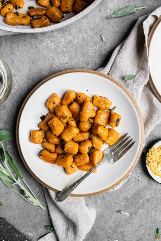 Vegan pumpkin gnocchi with sage butter sauce on a white plate with a fork and vegan parmesan cheese