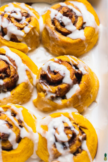 Close up shot of vegan pumpkin cinnamon rolls in a white dish with cream cheese icing drizzle by sweet simple vegan