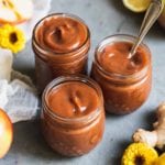 three jars filled with homemade apple butter surrounded by flowers apples and lemon by sweet simple vegan