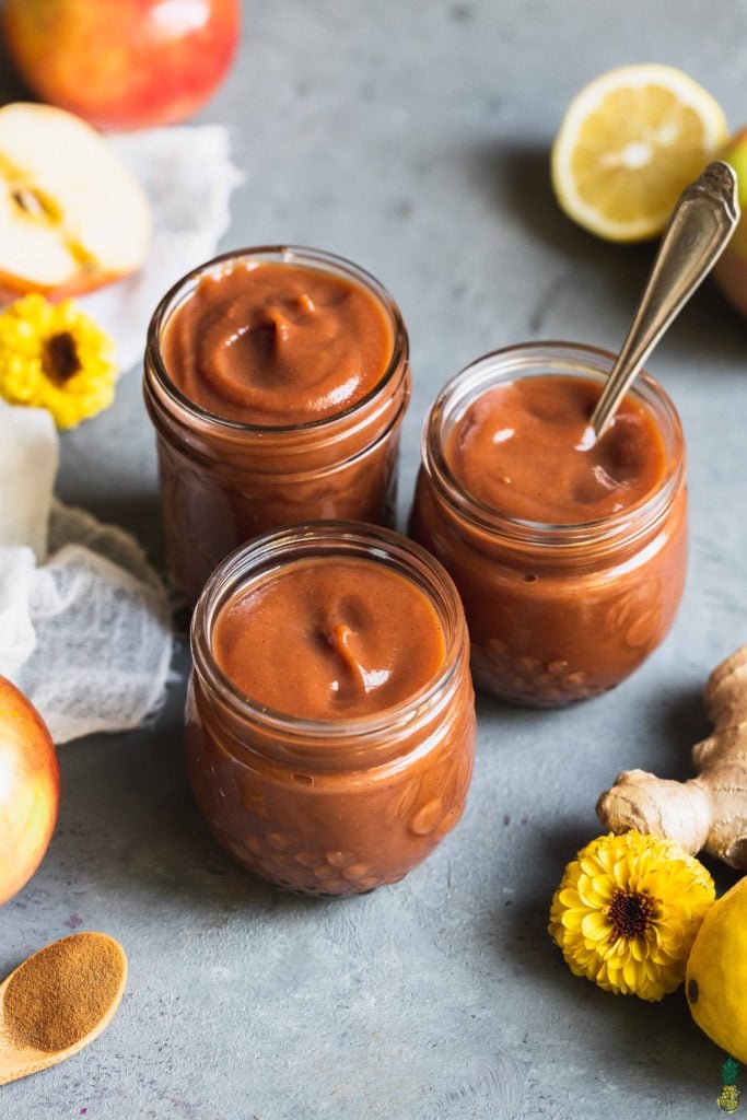 three jars filled with homemade apple butter surrounded by flowers apples and lemon by sweet simple vegan