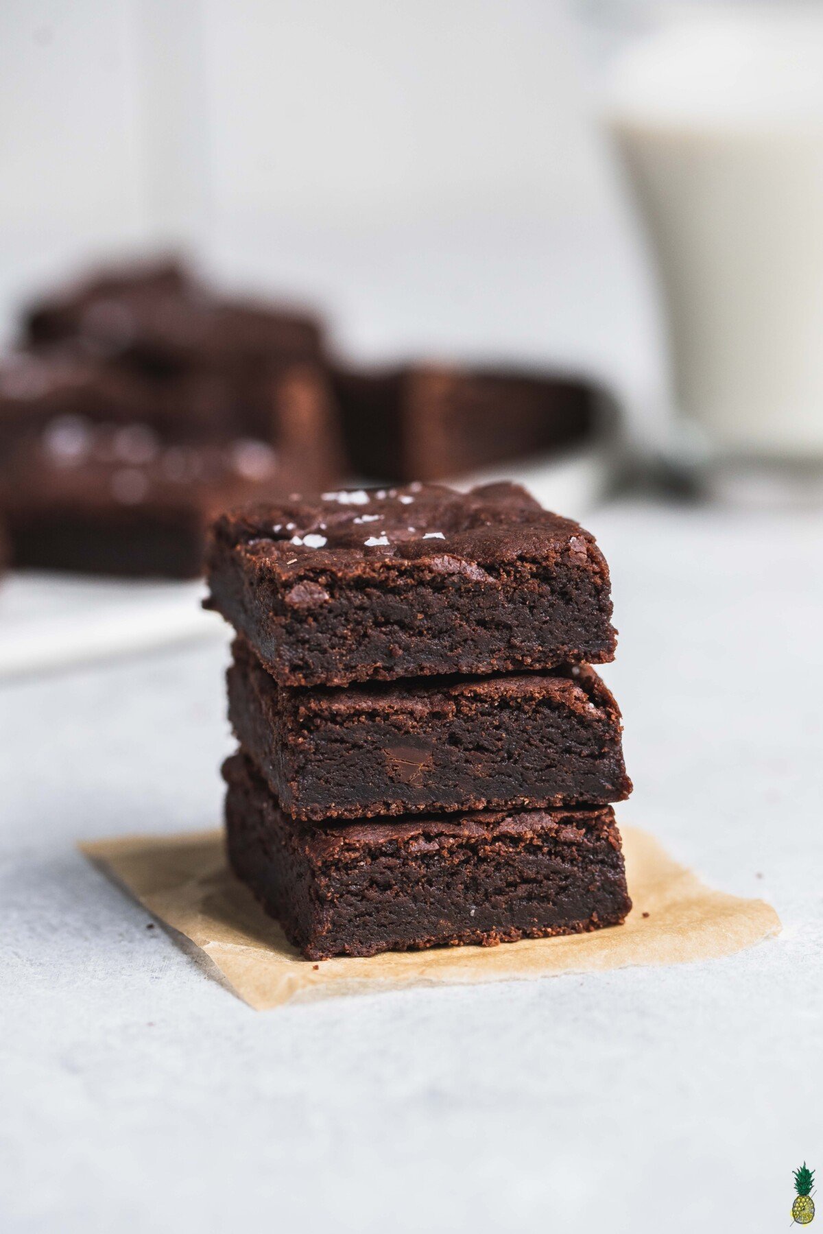 A stack of vegan brownies on a piece of parchment paper with brownies and almond milk in the background
