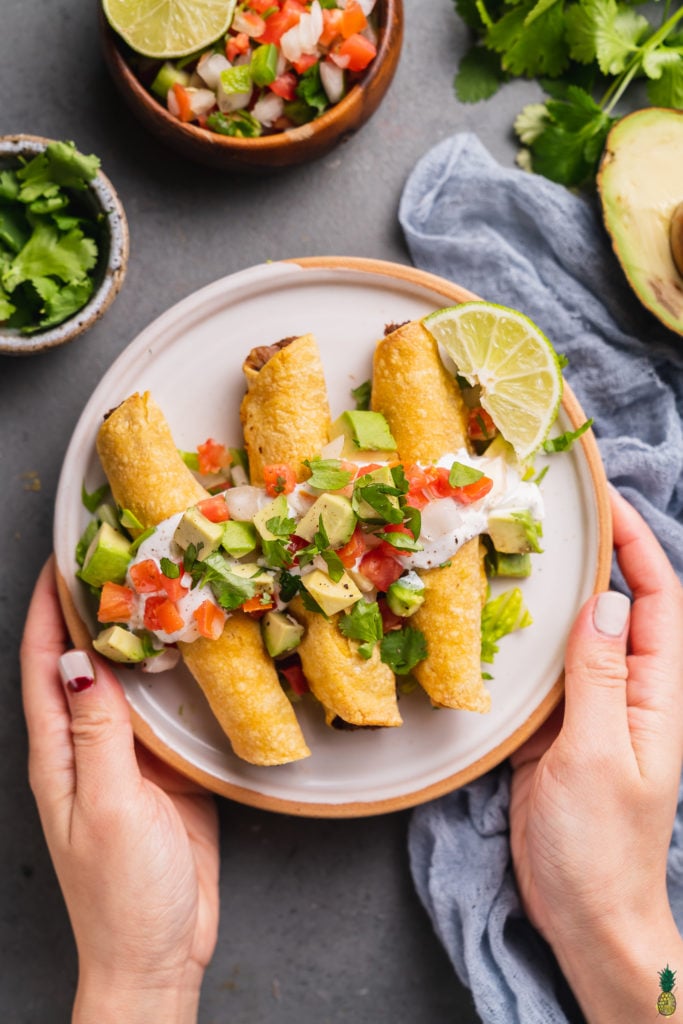 hands holding a plate of mushroom potato taquitos with vegan sour cream, pico de gallo, avocado and lime