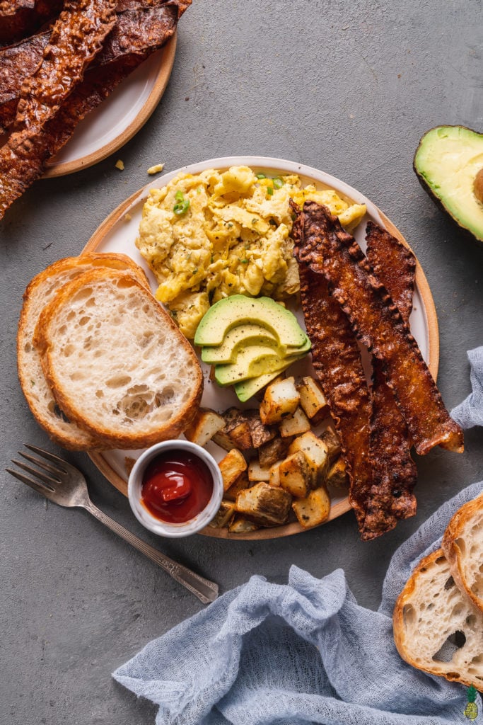 A vegan breakfast plate with crispy bacon, vegan eggs, avocado, breakfast potatoes, toast and ketchup by sweet simple vegan