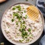 vegan french onion dip in a gray bowl covered in parsley with a potato chip dipped into it