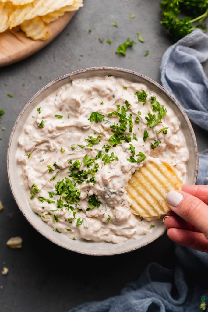 vegan french onion dip in a gray bowl covered in parsley with a potato chip dipped into it