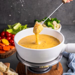 A photo of bread being dipped into vegan cheese fondue in a white pot surrounded by dippers by sweet simple vegan