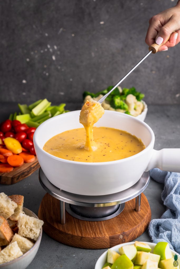 A photo of bread being dipped into vegan cheese fondue in a white pot surrounded by dippers by sweet simple vegan