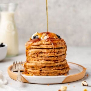 pouring maple syrup on stack of vegan carrot cake pancakes on white plate with raisins and whipped cream