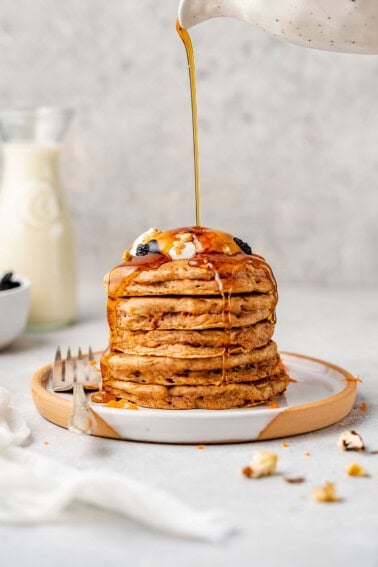 pouring maple syrup on stack of vegan carrot cake pancakes on white plate with raisins and whipped cream