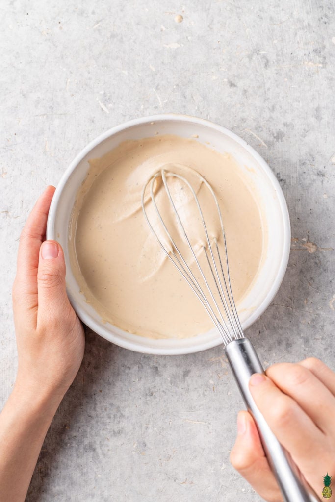 Mixing tahini dressing in a white bowl with a silver whisk by sweet simple vegan