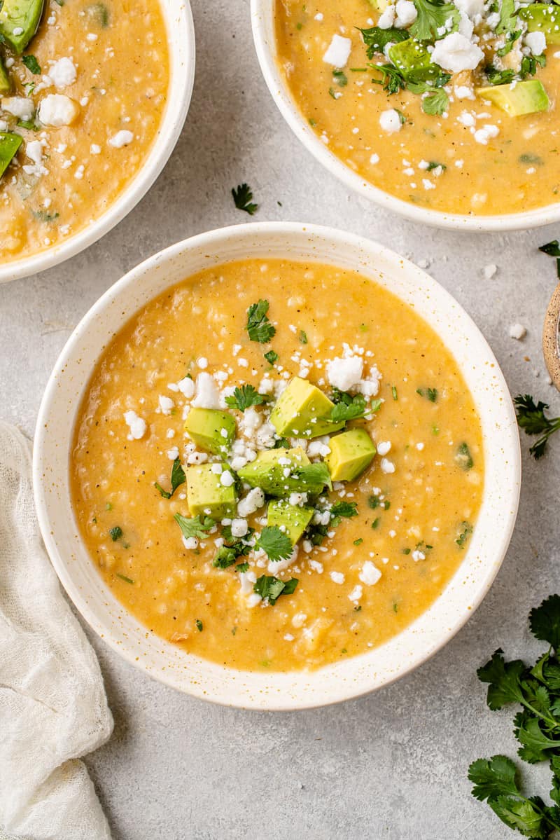 a close up of locro de papa with avocado, cilantro and vegan cheese on top in a white speckled bowl