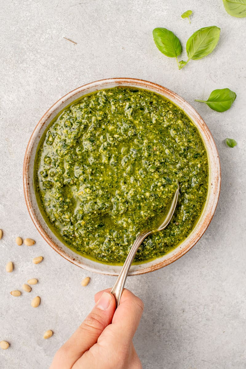 spoon scooping from a bowl of vegan pesto surrounded by pine nuts and basil