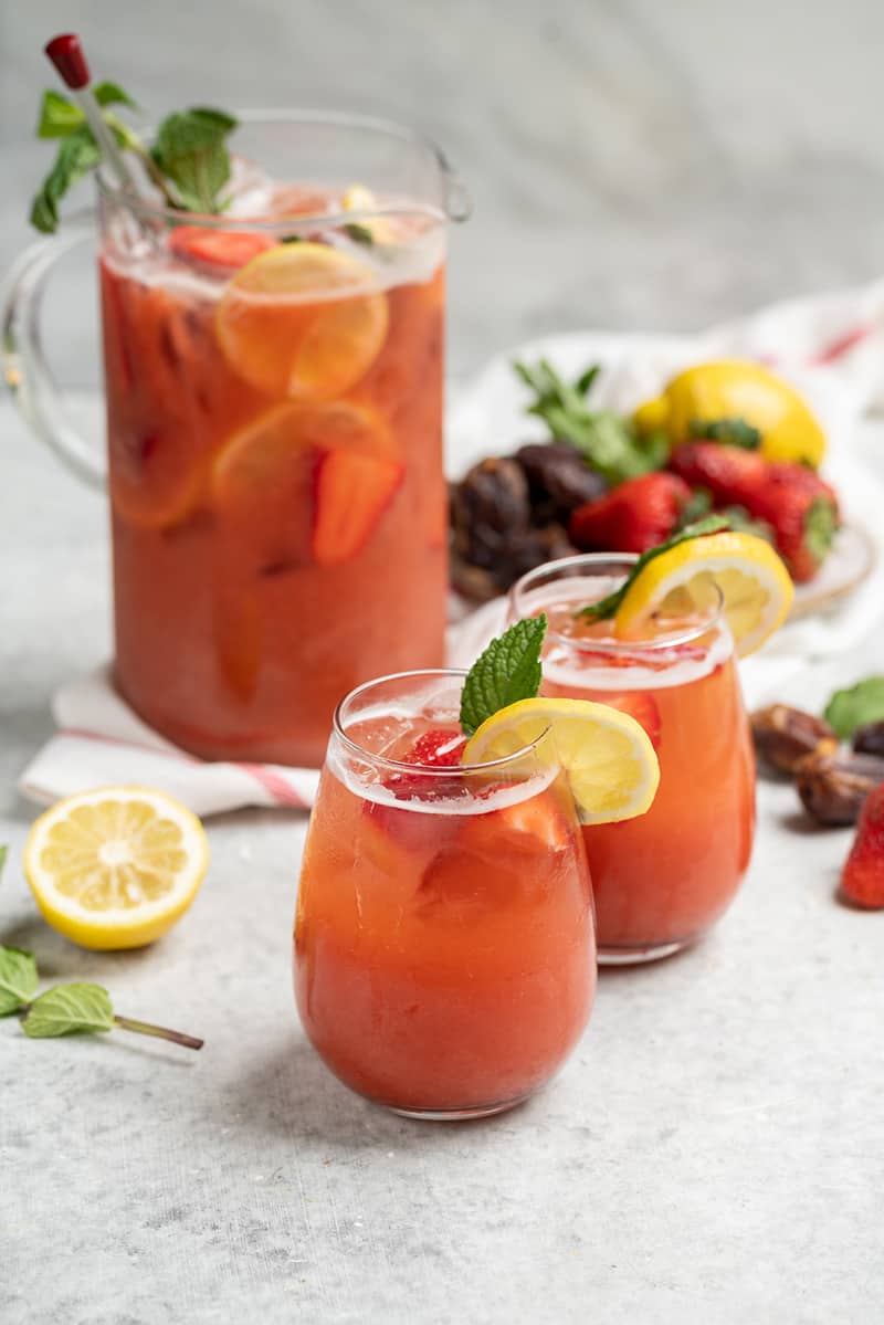 two glasses of strawberry lemonade on white table with full pitcher in the background