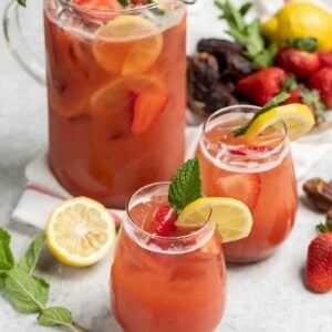 two glasses of strawberry lemonade on white table with full pitcher in the background