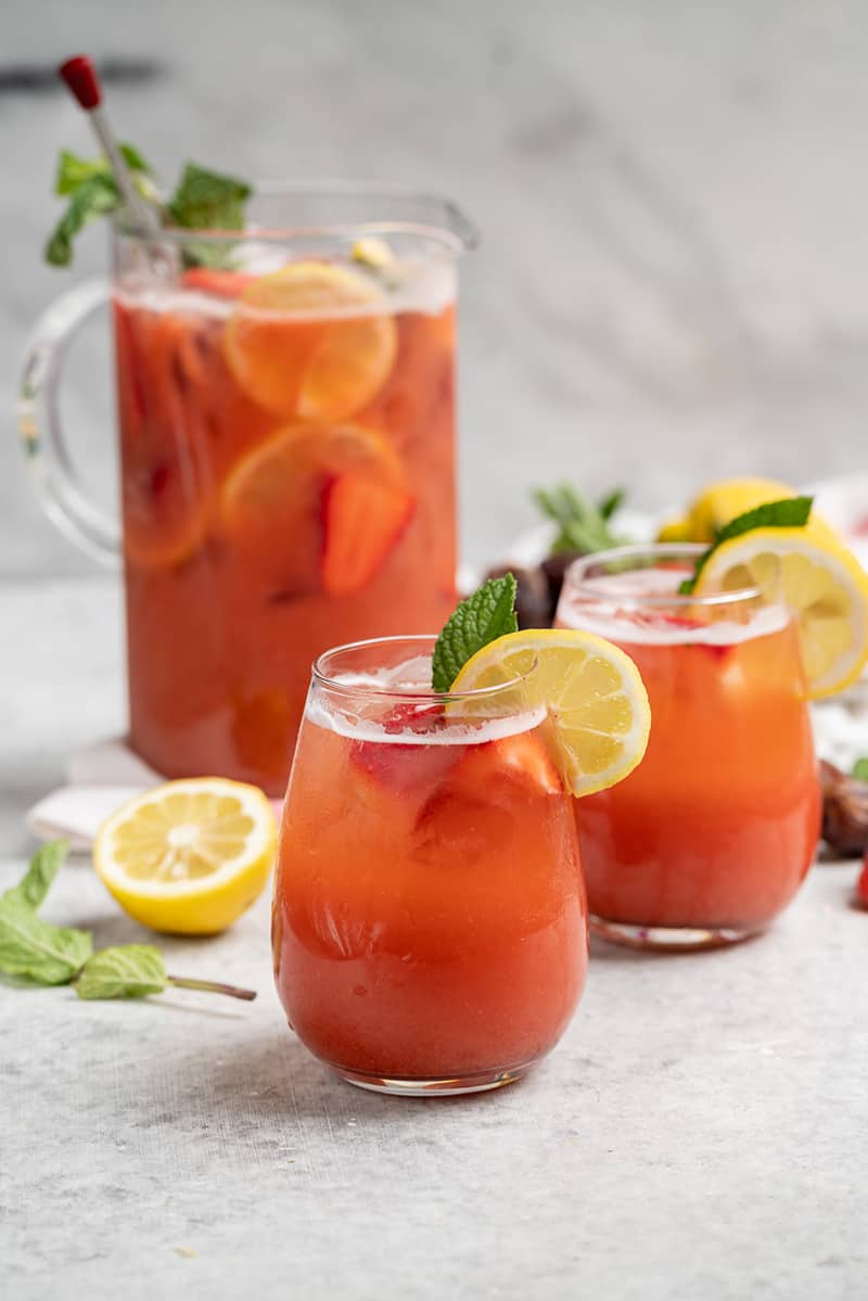 two glasses of strawberry lemonade on white table with full pitcher in the background