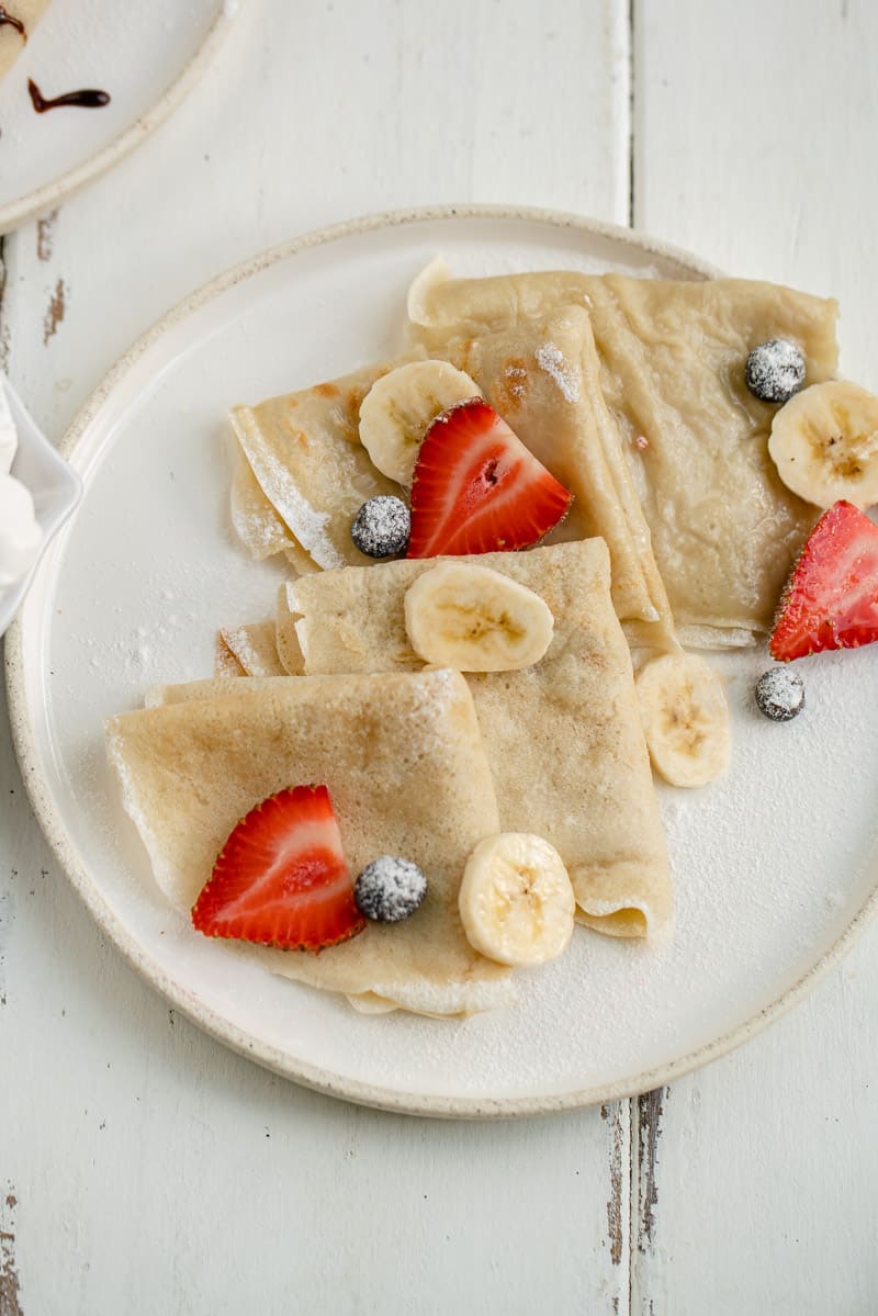 vegan crepes on a white plate with fresh fruit and powdered sugar