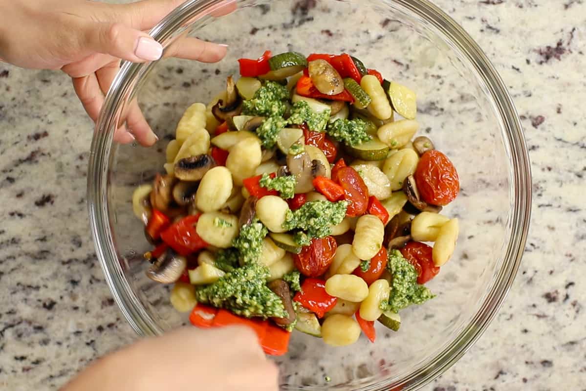 mixing pesto into gnocchi and vegetables in a glass bowl