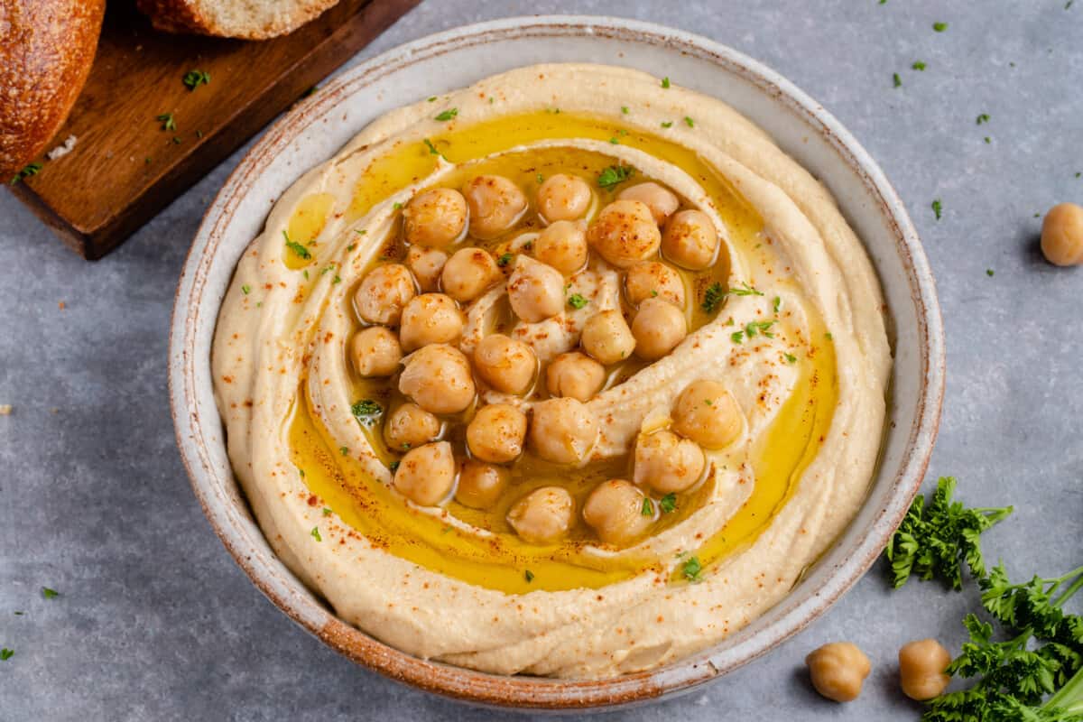 horizontal photo of creamy hummus in gray bowl topped with chickpeas, olive oil and parsley on gray background