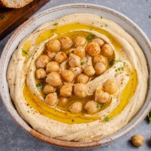 horizontal photo of creamy hummus in gray bowl topped with chickpeas, olive oil and parsley on gray background