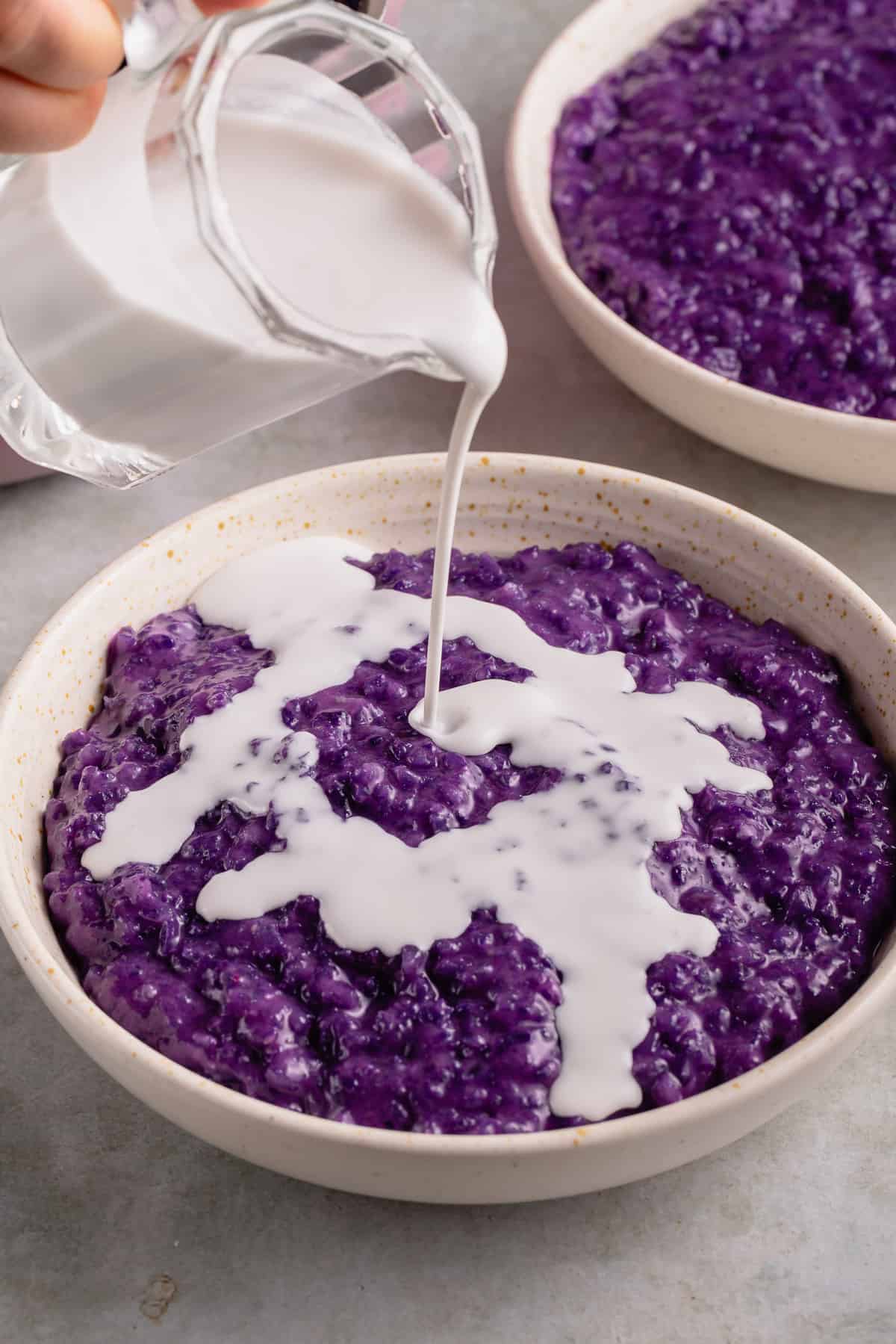 close up photo of pouring coconut milk on ube champorado from white bowl