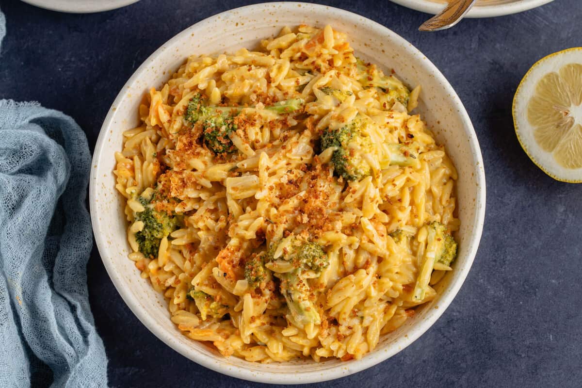 horizontal photo of broccoli cheddar orzo with breadcrumbs in beige bowl next to lemon half