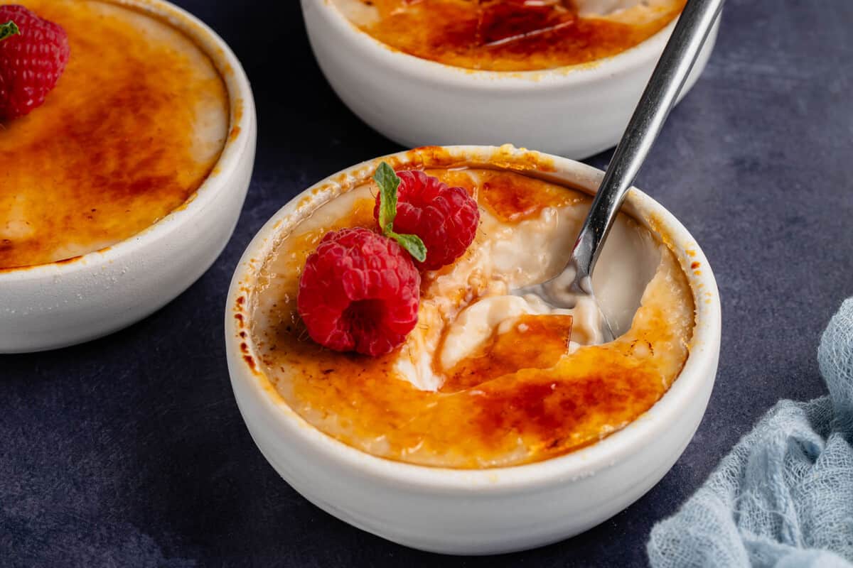horizontal photo of spoon in vegan creme brulee in white dish with raspberries on blue board