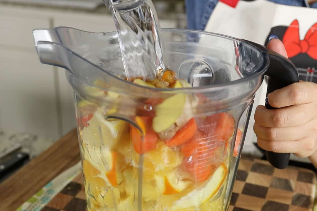 pouring water into a blender with oranges, lemons, pineapple, ginger, carrots, garlic, water, agave, turmeric and black pepper