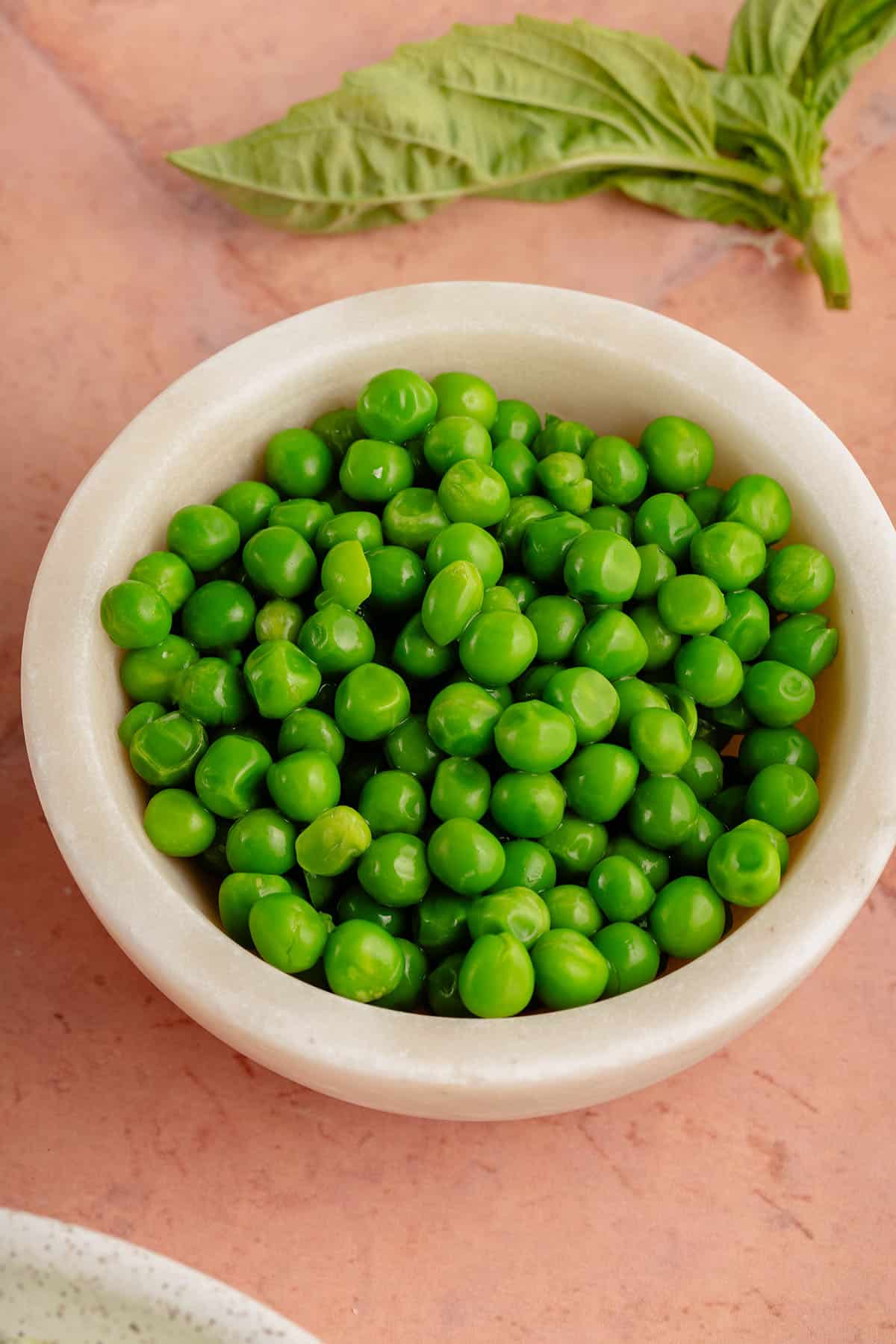 white bowl of fresh peas and a sprig of fresh basil on the side