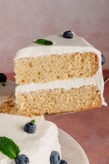 Slice of vegan vanilla cake with vanilla buttercream frosted decorated with blueberries and mint on a cake stand being lifted with a spatula