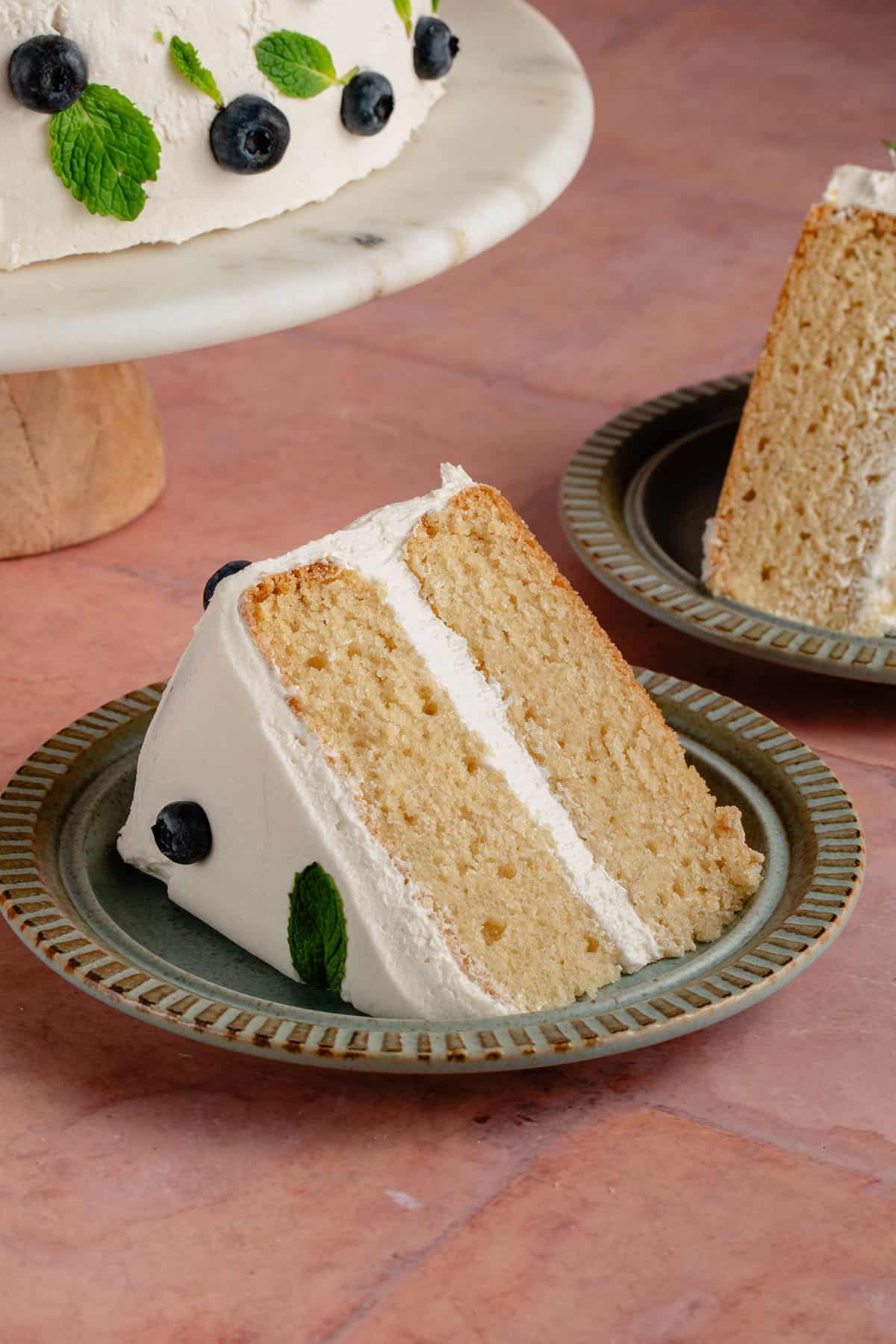 slice of vegan vanilla cake with buttercream frosting on a dark green plate with another slice of cake peaking in the background along with the full cake on the stand