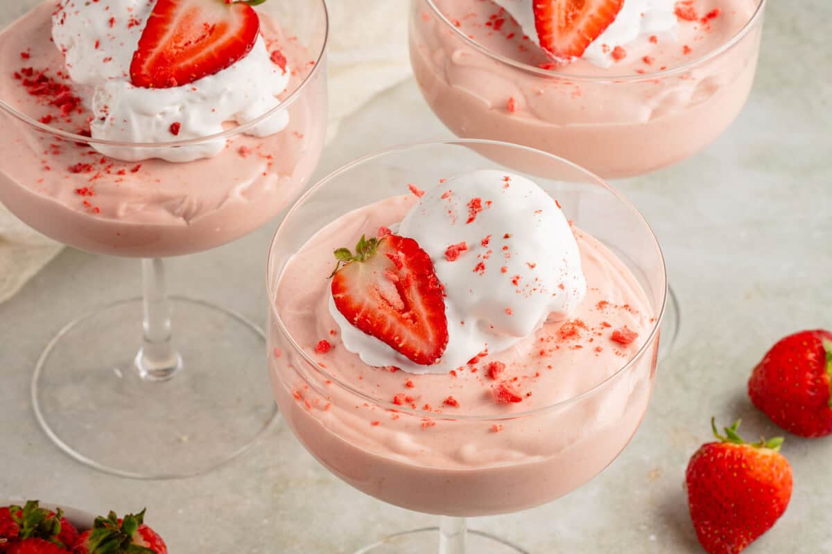 horizontal photo of 3 glasses of fluffy pink vegan strawberry mousse with whipped cream and fresh strawberries