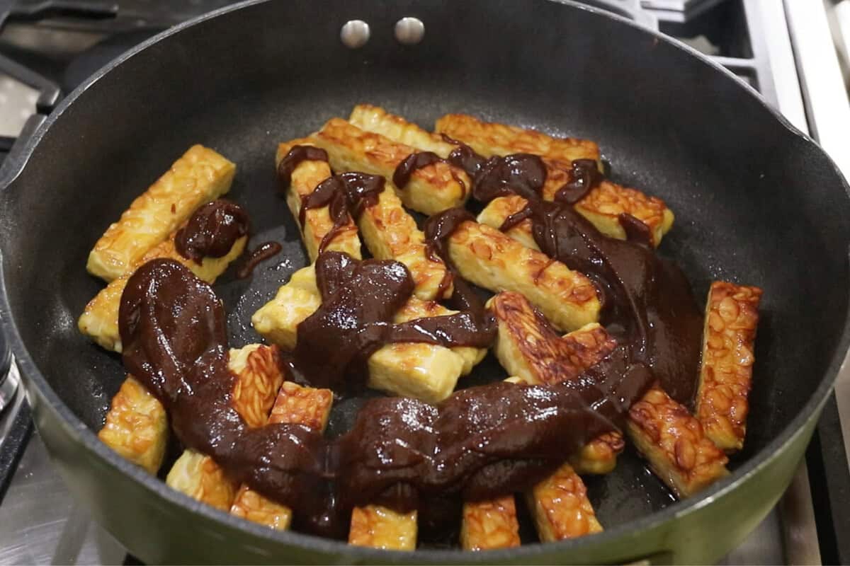 Pouring barbecue sauce over browned tempeh in pan