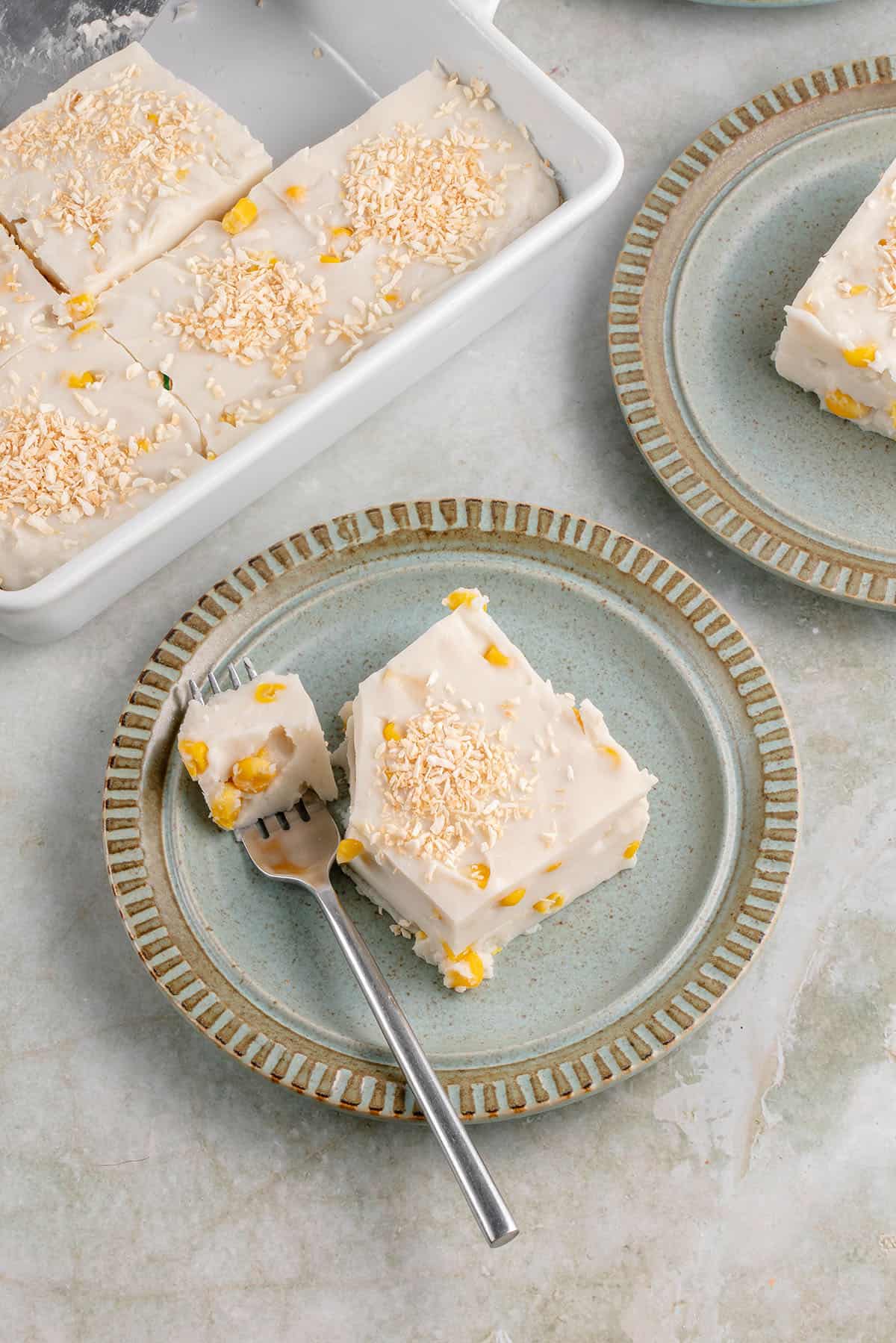 a slice of maja blanca on a blue plate with a fork and the tray peaking in from the top right corner