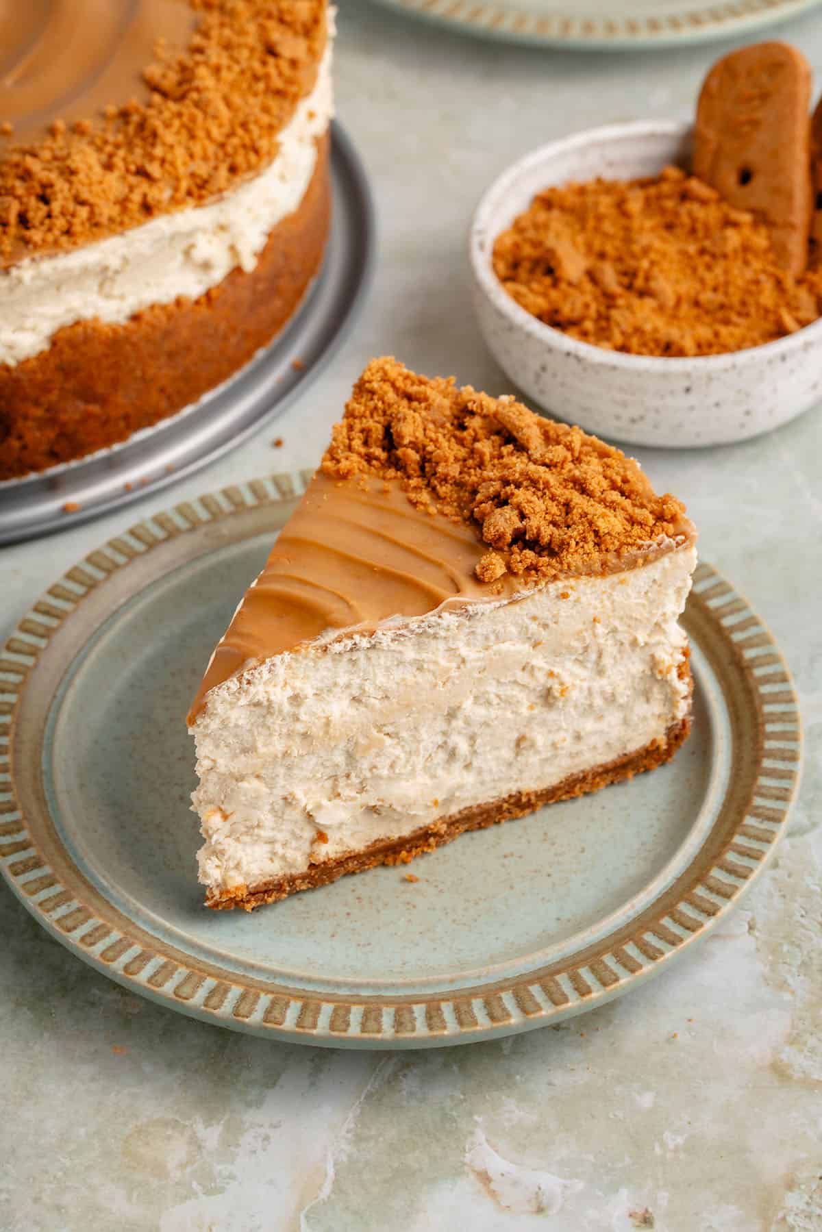 showing the front of a slice of biscoff cheesecake on a blue plate with the full cake and cookie crumbs in the background