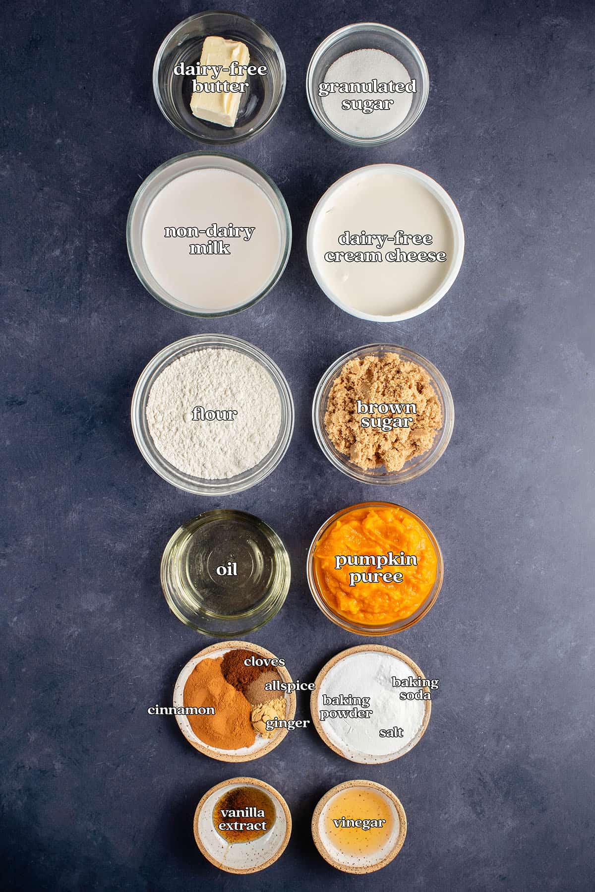 labeled ingredients for pumpkin cream cheese muffins: dairy-free butter, granulated sugar, non-dairy milk, dairy-free cream cheese, flour, brown sugar, oil, pumpkin puree, cinnamon, cloves, allspice, ginger, baking powder, baking soda, salt, vanilla and vinegar