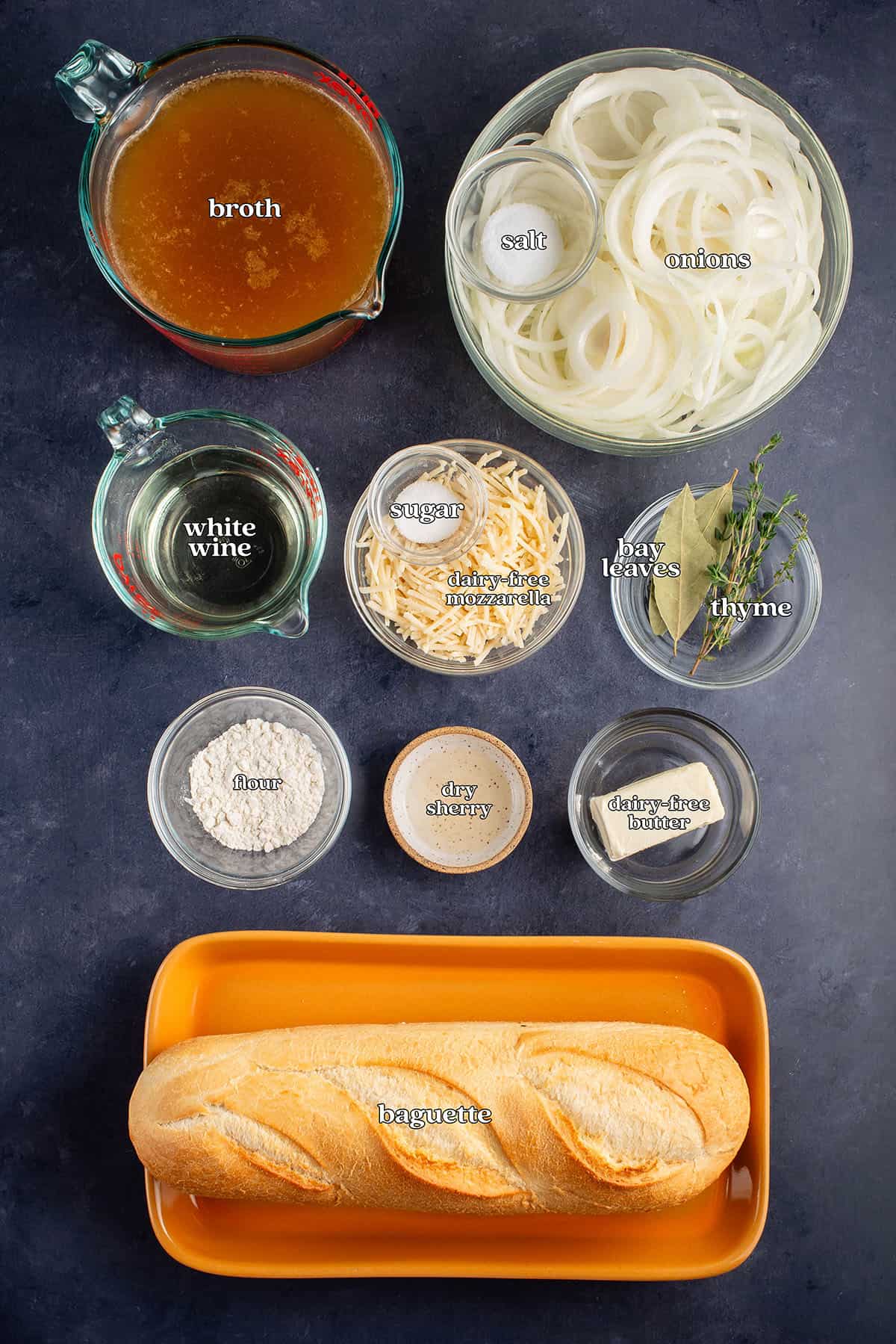 labeled ingredients for vegan french onion soup on blue table: broth, salt, onions, white wine, sugar, dairy-free mozzarella cheese, bay leaves, thyme, flour, dry sherry, dairy-free butter and baguette