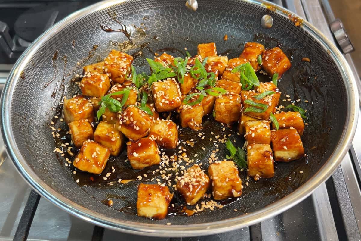 adding sesame seeds and sliced green onions to tofu in skillet