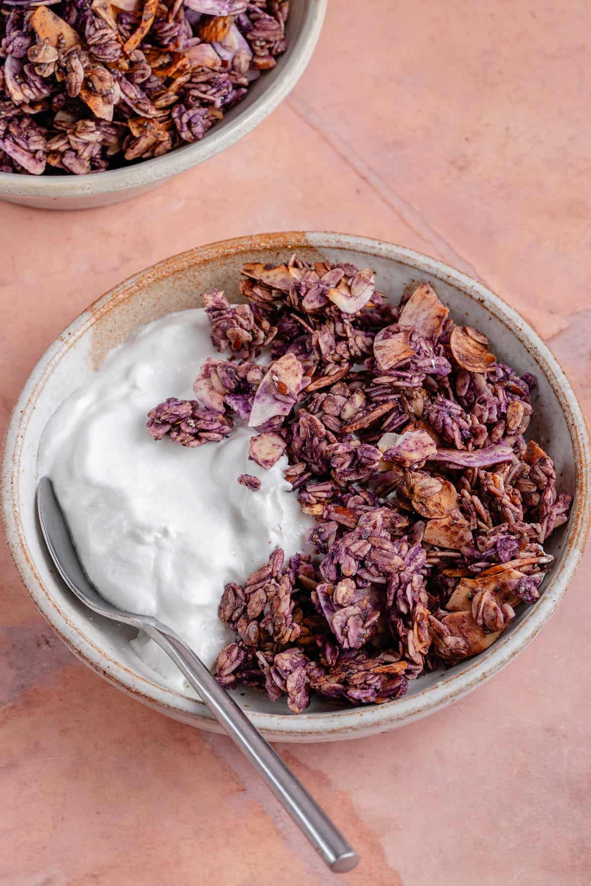 ube coconut granola in bowl with plan yogurt and spoon against pink backdrop