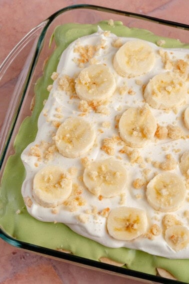 overhead photo of Vegan Matcha Banana Pudding in glass baking dish topped with whipped cream, crumbled vanilla wafers and banana slices