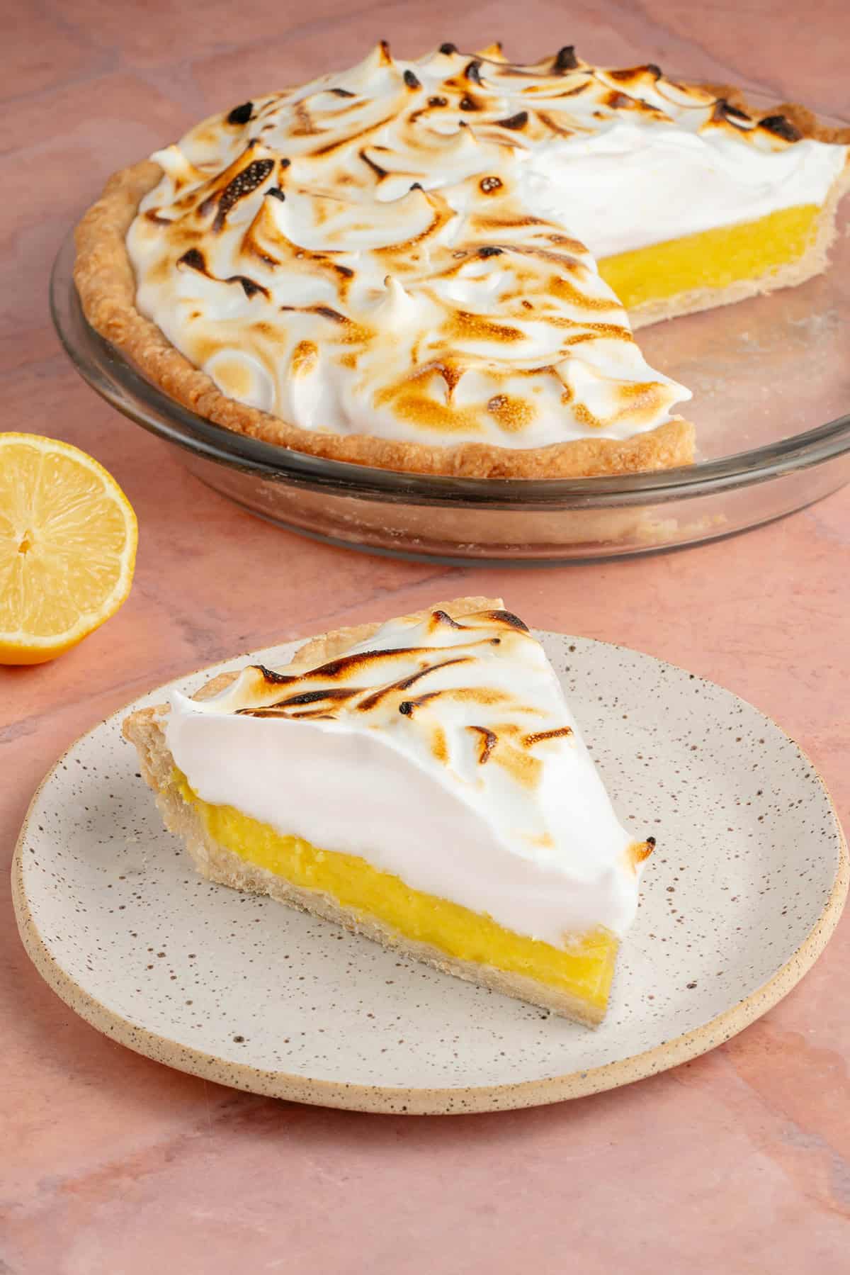 lemon meringue pie slice on small white plate with fresh lemon in front of cut pie in pan