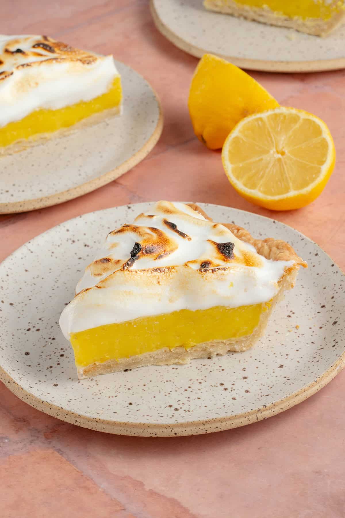lemon meringue pie slices on small white plates with fresh lemons on pink table