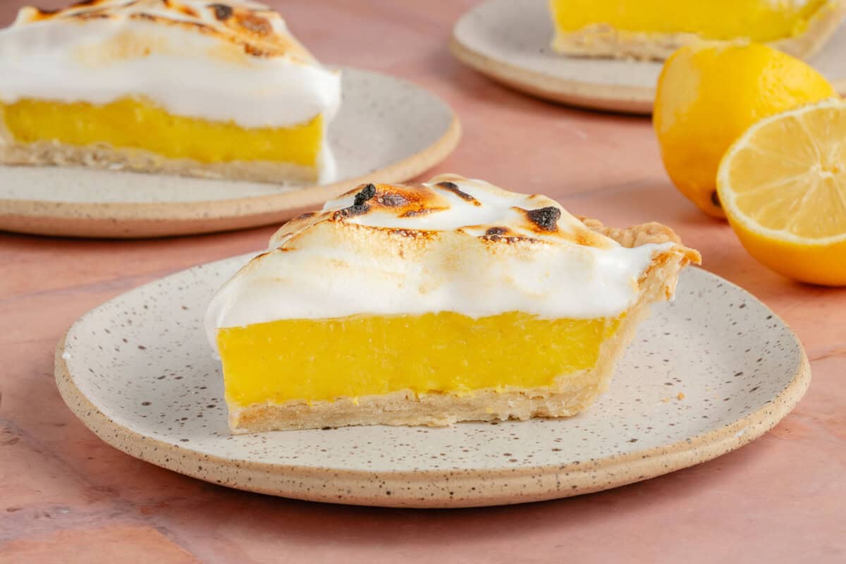 horizontal photo of 3 slices of lemon meringue pie on small white plates with fresh lemons on pink table