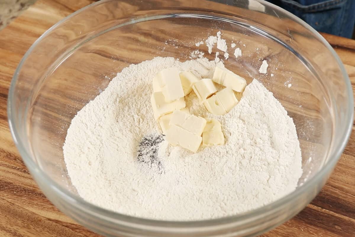 adding cubed butter to dry ingredients for pie crust in large glass bowl on wooden surface