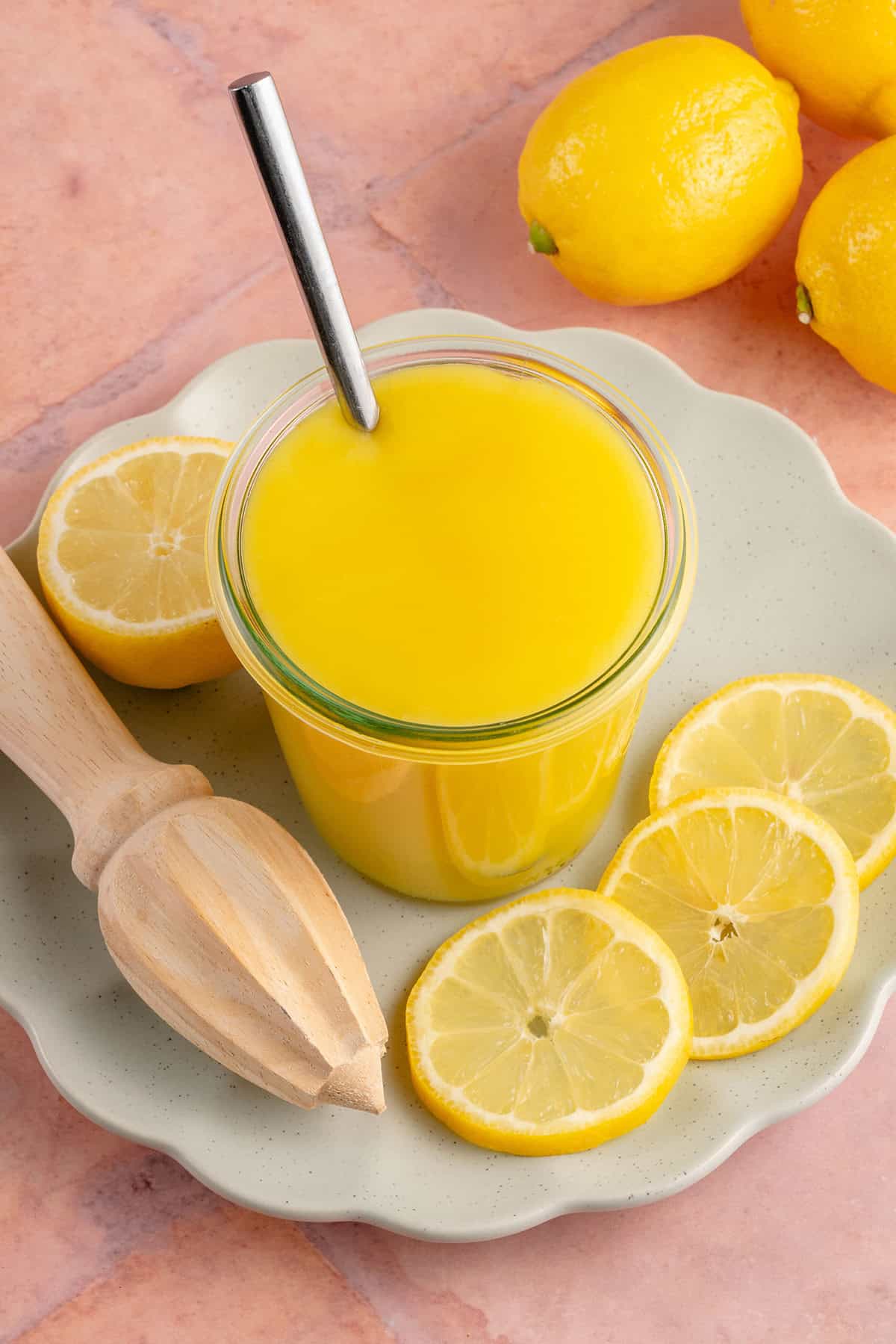 lemon curd in a jar with a spoon sticking out on a plate with a juicer and sliced lemons