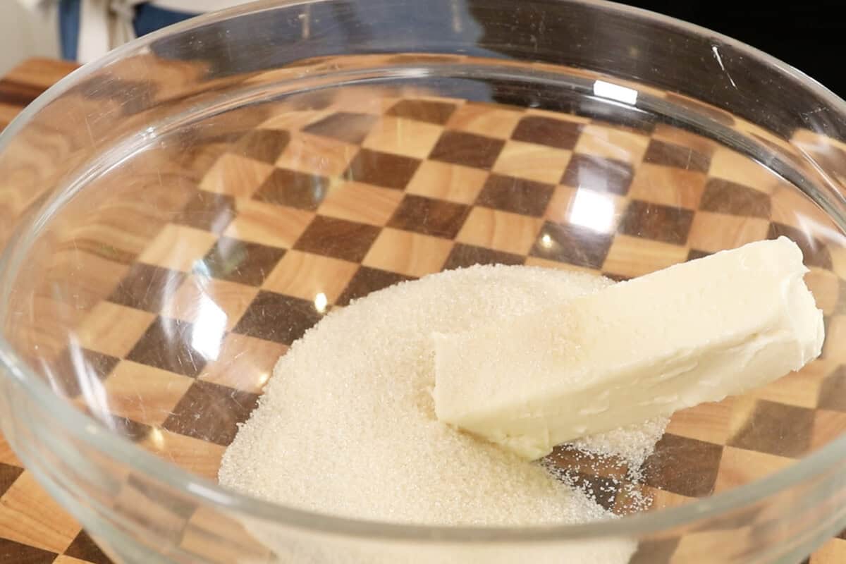 a stick of vegan butter and sugar in a large glass bowl on top of a checkered cutting board