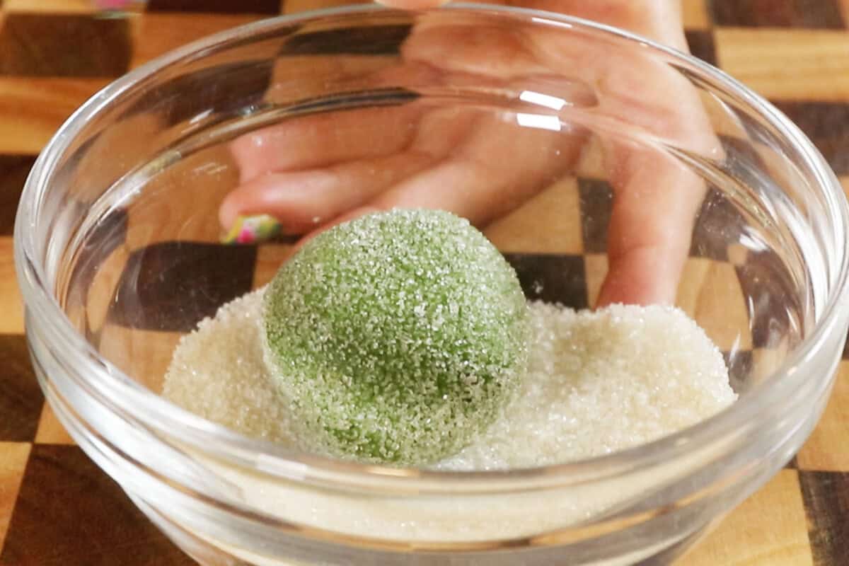rolling a ball of vegan matcha sugar cookie dough in a glass bowl of sugar
