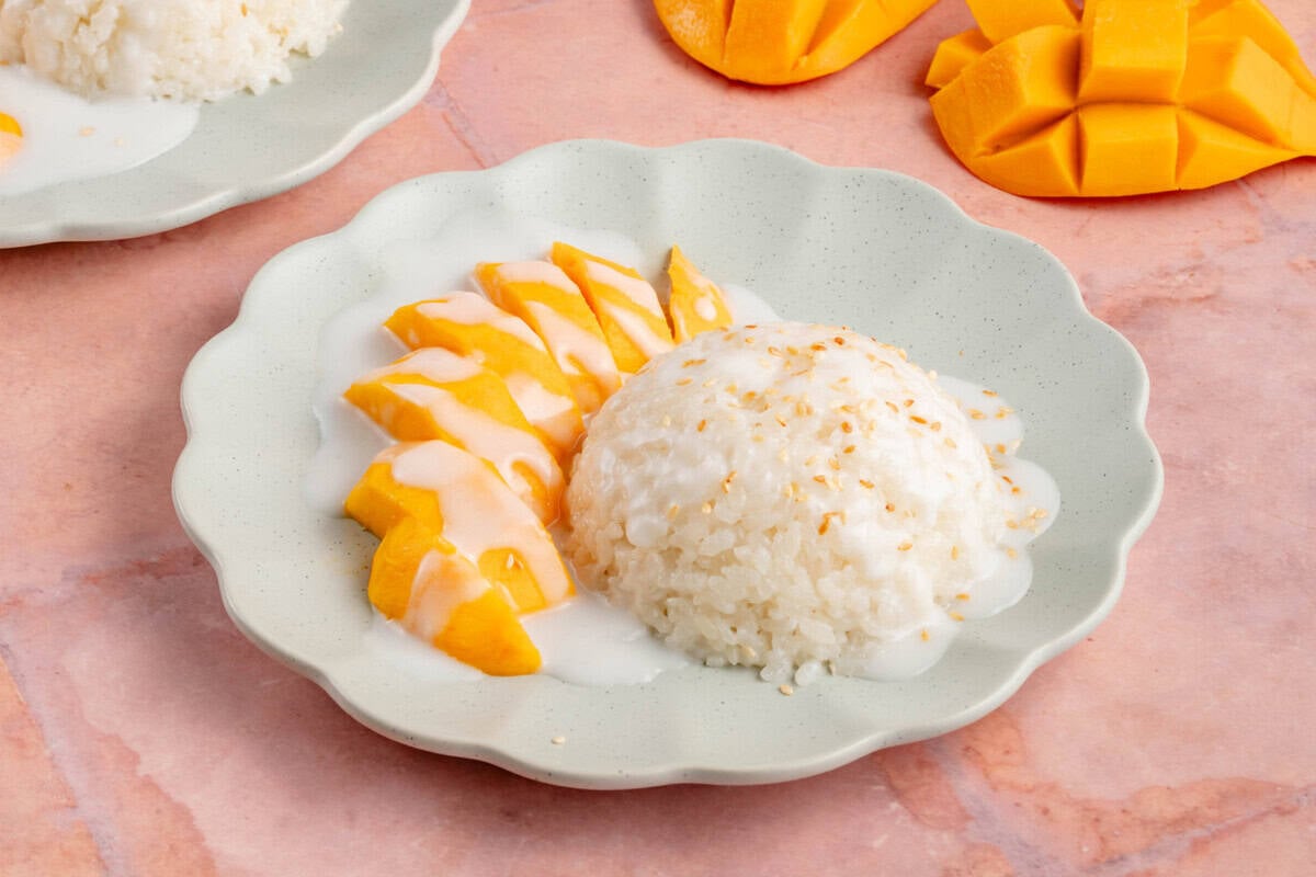 horizontal photo of mango sticky rice on a blue plate next to sliced mango on a pink table